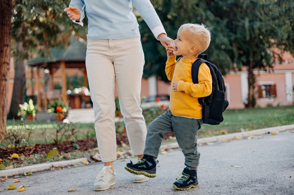 The photo captures a middle-aged woman and her young son holding hands as they walk along a city street, her toothy smile and his curious gaze symbolizing the warmth and guidance a mother offers on the road to education.